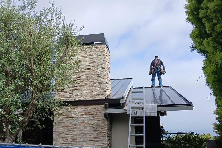 Roofer standing on a finished metal roof beside a stone chimney.